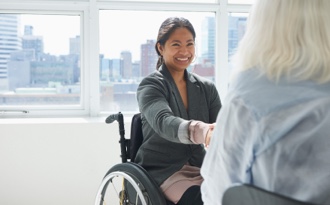 A female business professional in a wheelchair smiles as she shakes hands with a colleague in a corporate office setting.