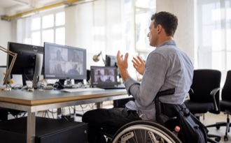 A business professional in a wheelchair is actively engaged in a video conference call at his desk in a corporate setting.