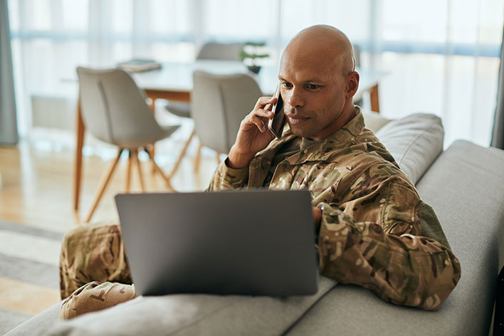 A male soldier in uniform sitting on a couch multitasks using a laptop while on the phone.