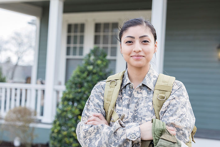 A young female soldier in uniform smiles as she poses in front of a house.