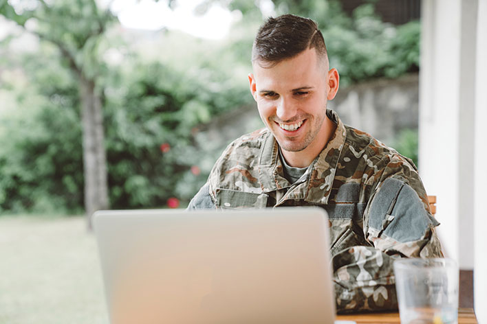 A male soldier in uniform sits at a desk, smiling, as he uses a laptop computer.