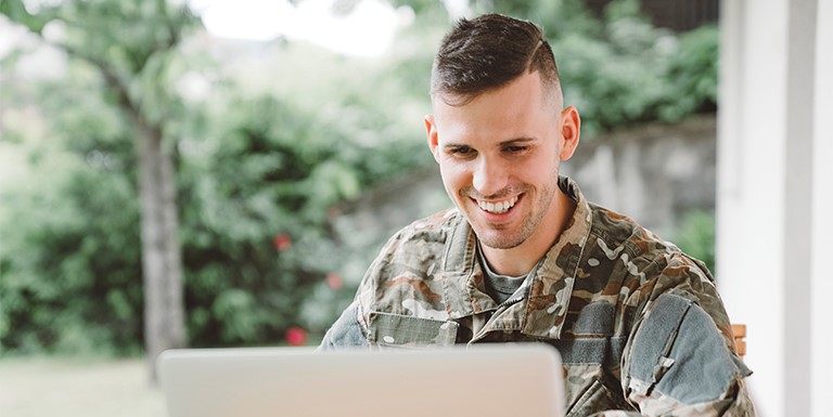 A male soldier in uniform sits at a desk, smiling, as he uses a laptop computer.