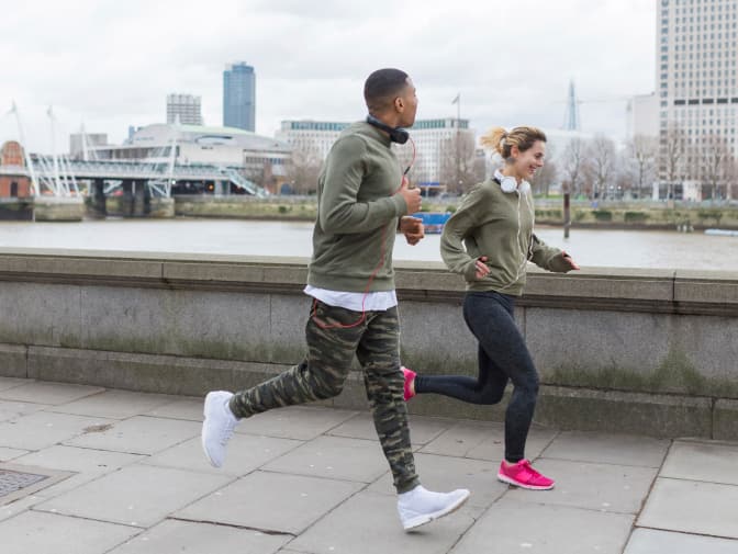 Two people are jogging along a paved path overlooking a river and cityscape